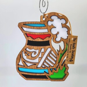 May include: Wooden ornament in the shape of a vase, featuring a layered design with blue, red, black, and white accents. The ornament has a natural wood grain finish and a metal hanger. A small tag reads "The Head's Creation Made in Albuquerque N.M."