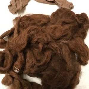May include: A pile of dark brown, curly hair, possibly for a costume or craft project, with a tan nylon stocking laid across the top. A small, white metal clip is visible.