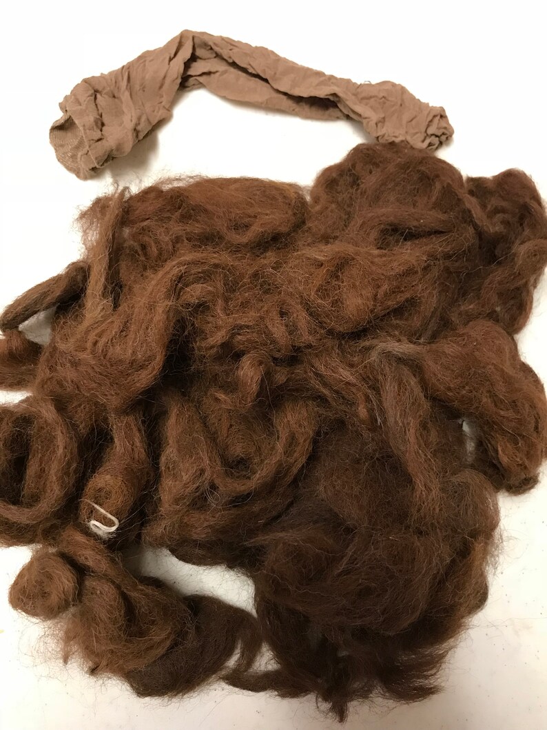 May include: A pile of dark brown, curly hair, possibly for a costume or craft project, with a tan nylon stocking laid across the top. A small, white metal clip is visible.