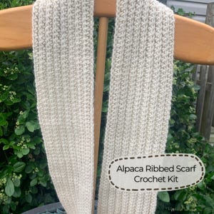 May include: A cream-colored ribbed scarf with a vertical knit texture. A white label with a brown dashed border is attached to the lower right side, featuring the black text: Alpaca Ribbed Scarf Crochet Kit. The scarf is draped over a wooden hanger.