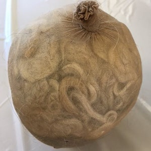 May include: A beige mesh bag filled with what appears to be hair or fiber. The bag is gathered at the top and tied, forming a small, knotted closure. The contents are visible through the mesh.