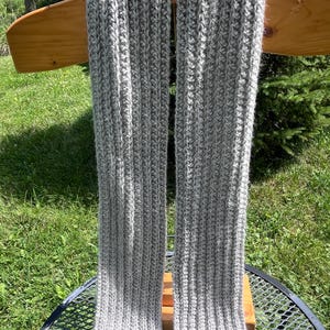 May include: A long, light gray scarf featuring a vertical ribbed knit pattern. The scarf is constructed with a textured yarn that displays a consistent, repeating stitch design throughout its length, creating a soft and uniform appearance.