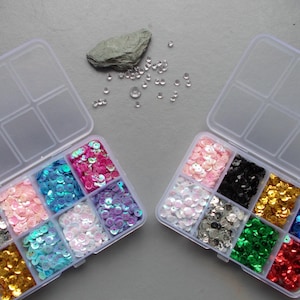 May include: Two clear plastic organiser boxes filled with colourful sequins. The sequins are in various colours, including silver, pink, blue, purple, gold, black, green, and red. A small grey rock and scattered clear beads are also present.