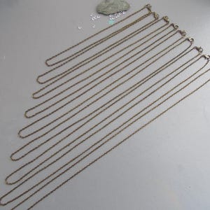 May include: A selection of bronze-toned chain necklaces with lobster claw clasps, displayed diagonally on a pale grey surface. The chains are fine and delicate, ideal for pendants or charms.