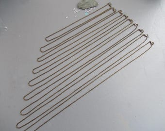 May include: A selection of bronze-toned chain necklaces with lobster claw clasps, displayed diagonally on a pale grey surface. The chains are fine and delicate, ideal for pendants or charms.