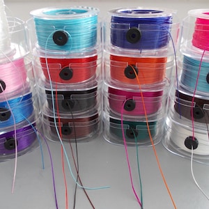 May include: A selection of colourful elastic string spools in clear plastic containers. The spools are stacked, displaying shades such as pink, blue, red, and black. Strands of the elastic extend from the spools, indicating their use in crafts or jewellery making.