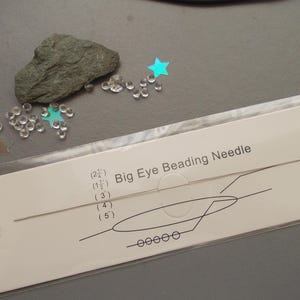 May include: A packet containing a Big Eye Beading Needle, with a diagram and measurements. The image also shows a grey rock, clear beads, and star-shaped sequins on a grey surface. The needle is designed for beading projects.
