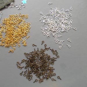 May include: A selection of small eye pins in gold, silver, and bronze hues. These miniature metal findings are arranged on a grey surface, suitable for jewellery making and craft projects. The pins feature a loop at one end and a pointed tip.