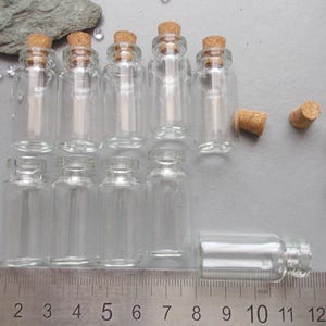 May include: A selection of small, clear glass bottles with cork stoppers. The bottles are cylindrical with a narrow neck and a rounded base. A ruler is placed below the bottles, showing measurements in millimeters.
