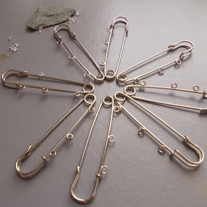 May include: A collection of gold-coloured safety pins arranged in a circular pattern. Each pin has a looped end and small decorative rings. The pins are on a grey surface, with small decorative elements scattered around.