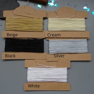 May include: A selection of elastic cord spools in beige, cream, black, silver, and white. Each spool is labelled with its colour. The cords are wound on cardboard spools, suitable for various craft projects.