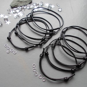 May include: A collection of black cord bracelets with adjustable knots, alongside small silver rings, star-shaped sequins, and clear crystals. The bracelets are presented on a grey surface. The bracelets appear to be handmade.
