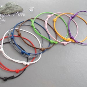 May include: A selection of adjustable string bracelets in a range of colours: black, red, white, blue, brown, green, pink, yellow, and purple. Each bracelet has a sliding knot closure for a custom fit. The bracelets are shown on a grey surface.
