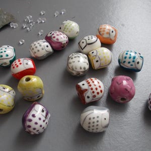 May include: A selection of ceramic owl beads in a variety of colours, including white, yellow, red, purple, and blue. Each bead features a unique owl design with dotted patterns. The beads are scattered on a grey surface.