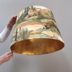 Pode incluir: Um abajur em forma de tambor com uma paisagem impressa de colinas, árvores e edifícios em tons de terra. O interior do abajur é revestido com um acabamento metálico dourado refletor e inclui uma estrutura central de metal.