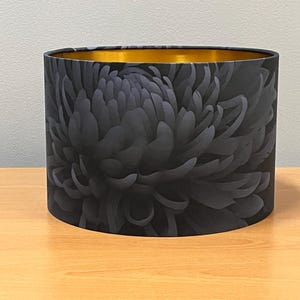 Puede incluir: Pantalla de lámpara cilíndrica con un gran diseño floral gris oscuro. El interior de la pantalla es dorado. La pantalla de la lámpara está sobre una superficie de madera.