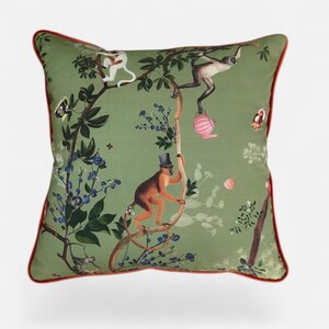 Può includere: Un cuscino quadrato con sfondo verde salvia caratterizzato da una stampa di scimmie che si arrampicano sui rami degli alberi. Una scimmia indossa un cilindro, mentre un'altra tiene una teiera rosa. Il design include fiori blu, foglie verdi e un bordo rosso.
