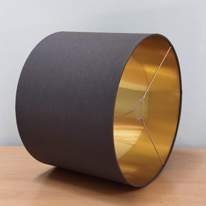 May include: A dark grey lampshade with a gold-coloured interior. The lampshade is cylindrical in shape and made from fabric. It is designed for use with a table or floor lamp. The diameter is approximately 30 cm.