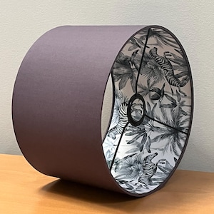 Pode incluir: Abajur cilíndrico com exterior roxo escuro. O interior apresenta uma cena de selva em preto e branco com zebras e folhagens tropicais. O abajur foi projetado para caber em uma luminária e fornecer iluminação ambiente.