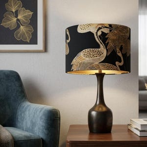 Może przedstawiać: Dekoracyjna lampa stołowa z czarnym abażurem z złotym wzorem żurawia. Lampa ma ciemnobrązową podstawę i stoi na drewnianym stole. Na ścianie wisi oprawiony złoty nadruk liści.