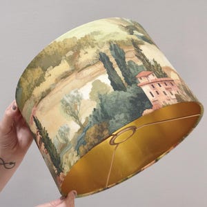 Pode incluir: Um abajur cilíndrico com uma paisagem impressa que apresenta árvores verdes, colinas e um edifício de vários andares com telhado vermelho. O interior do abajur é revestido com um acabamento dourado metálico e inclui uma estrutura de metal.