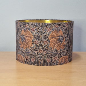 Puede incluir: Pantalla de lámpara cilíndrica con un patrón floral en tonos marrón, azul y rojo. El interior es dorado. La pantalla de la lámpara está sobre una superficie de madera.