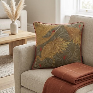 Peut inclure: Un coussin carré avec un fond vert olive foncé orné d'un motif répétitif de grues dorées et d'éventails, agrémenté de petits détails rouges et d'un passepoil rouge, accompagné d'un plaid plié à carreaux de couleur rouille.
