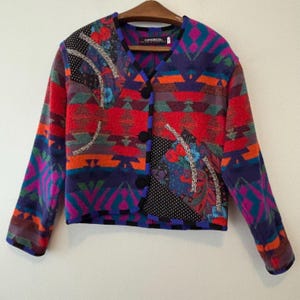 Könnte beinhalten: Eine farbenfrohe Jacke mit Muster, V-Ausschnitt und schwarzen Knöpfen. Die Jacke zeigt eine Mischung aus geometrischen und floralen Designs in Rot, Lila, Orange und Türkis. Die Ärmel sind lang und die Jacke hängt an einem Holzbügel.