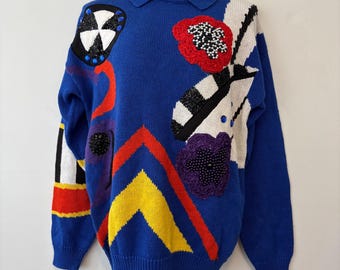May include: A blue knitted sweater with a collared neckline and long sleeves. The sweater is decorated with colourful abstract patterns, including red, yellow, black, white, and purple shapes, and beaded floral accents. The sweater is displayed on a mannequin.