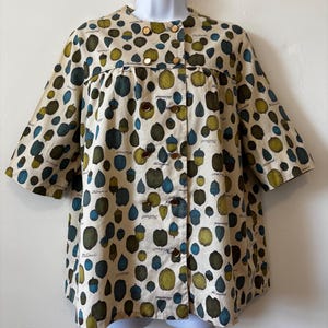 May include: A beige, short-sleeved, double-breasted jacket featuring a repeating pattern of green and blue nuts and acorns. The garment has a round neckline, a horizontal seam across the chest, and six metallic buttons down the front closure.