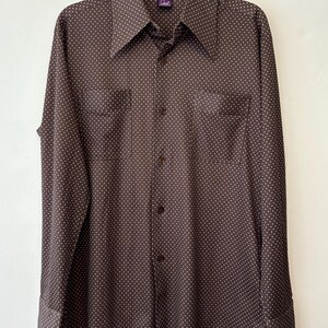 Puede incluir: Una camisa marrón con lunares blancos. La camisa de manga larga tiene dos bolsillos en el pecho y un cuello ancho. La camisa está colgada de una percha negra.
