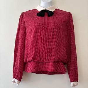 Peut inclure: Un chemisier rouge à motif quadrillé blanc, avec un col blanc et un nœud noir. Les manches longues ont des poignets blancs. Le chemisier est présenté sur un mannequin.