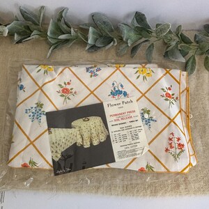 Puede incluir: Un mantel nuevo y empaquetado con un estampado floral. La tela blanca presenta un diseño de cuadrícula con líneas amarillas y racimos de flores de colores. El embalaje incluye una etiqueta del producto con el texto "Flower Patch".