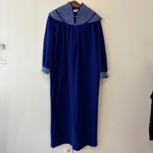 Puede incluir: Una bata larga azul real con cuello y puños estampados con flores. La bata tiene mangas largas y una cremallera de largo completo. El cuello y los puños presentan un diseño floral azul y blanco. La bata está hecha de un material suave y afelpado.