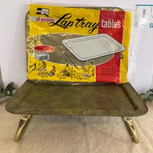 May include: A vintage green and gold lap tray with folding legs. The tray has a decorative floral pattern and gold-colored legs. A packaged "Quaker" brand lap tray is in the background, with the words "Lap tray tables" printed on the packaging.