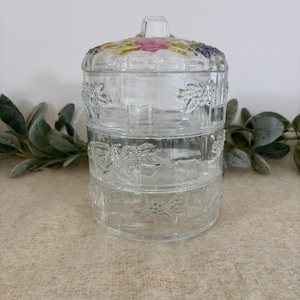 Pode incluir: Um prato de servir de vidro transparente de três camadas com tampa. A tampa é decorada com detalhes florais coloridos. As laterais do prato apresentam desenhos florais em relevo. O prato foi projetado para conter e exibir itens pequenos.