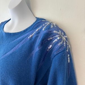 Pode incluir: Um suéter azul com decote redondo e detalhes decorativos de lantejoulas e fita nos ombros. O suéter tem um design de malha texturizada e uma cor azul sólida.