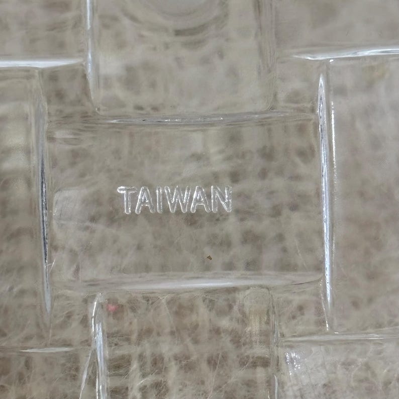 Może przedstawiać: Zbliżenie przezroczystego szklanego przedmiotu z wzorem kratki. Słowo "TAIWAN" jest wytrawione w szkle. Tło ma miękki, neutralny odcień.