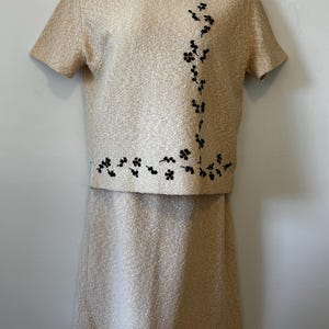 May include: A two-piece set consisting of a short-sleeved top and a skirt in a textured gold fabric. The top features a round neckline and an L-shaped pattern of small, black embroidered floral motifs along the right side and bottom hem.