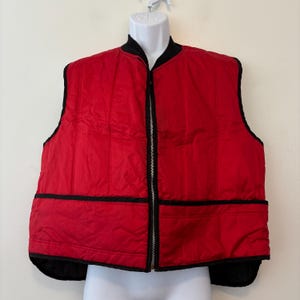 Może przedstawiać: Czerwona pikowana kamizelka z czarnym zamkiem błyskawicznym i czarnymi lamówkami. Kamizelka ma pikowany wzór i czarny kołnierz.