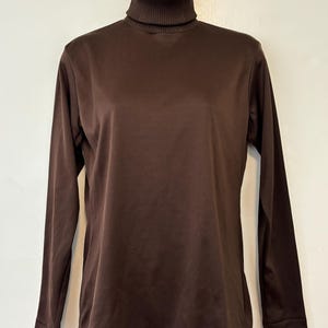 Puede incluir: Una camisa de cuello alto marrón de manga larga. La camisa está hecha de una tela lisa y presenta un cuello alto acanalado. La prenda se exhibe en un maniquí blanco, mostrando su forma y diseño. La camisa es de color liso y tiene un estilo clásico y sencillo.