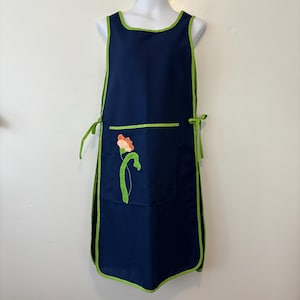 Pode incluir: Um avental azul marinho com acabamento verde e um bolso com uma aplicação floral. O avental tem laços verdes na cintura e uma borda verde ao redor da gola e cavas. A aplicação floral é laranja e verde.