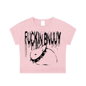 May include: A light pink short-sleeved crop top featuring a black graphic print of a rabbit wearing a spiked collar. Above the rabbit, the text "FUCKIN BNUUY" is printed in a distressed, dripping black font style across the centre of the shirt.