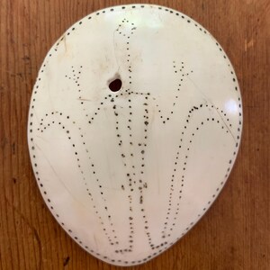 May include: Oval-shaped, off-white pendant with a central hole and a dotted design. The design features a series of curved lines and dots, creating a unique pattern. The pendant is likely made of a natural material.