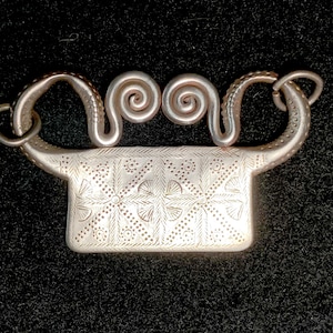 May include: A silver pendant with a rectangular shape and intricate floral designs. The pendant has a unique design with two spiral loops at the top.