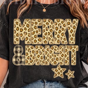 May include: A charcoal grey mineral-washed t-shirt featuring the text MERRY & BRIGHT in a leopard print pattern, with the ampersand in a black and white buffalo plaid pattern. Two leopard print stars are positioned below the text.