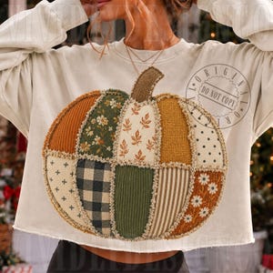 May include: A cream-coloured long-sleeve sweatshirt featuring a large, central pumpkin graphic composed of various fabric patches. The pumpkin design includes segments of corduroy, plaid, floral, polka dot, and striped patterns in autumn colours.