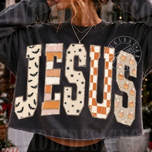 May include: A charcoal grey long-sleeve sweatshirt featuring the word JESUS in large block letters. Each letter has a unique pattern: bats, orange and white stripes, black stars on cream, orange and white checkers, and candy corn shapes.