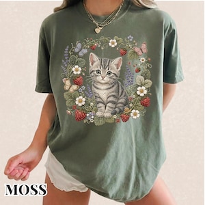 May include: A moss green short-sleeve t-shirt featuring a central graphic of a grey tabby kitten surrounded by a circular wreath of red strawberries, white flowers, lavender sprigs, and butterflies. The shirt has a crew neckline and a relaxed fit.