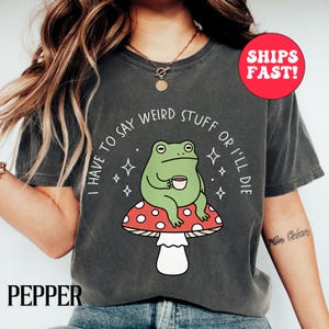May include: A charcoal gray t-shirt featuring a graphic of a green frog sitting on a red and white spotted mushroom while holding a small cup. The text "I HAVE TO SAY WEIRD STUFF OR I'LL DIE" is printed in white around the frog with small star accents.